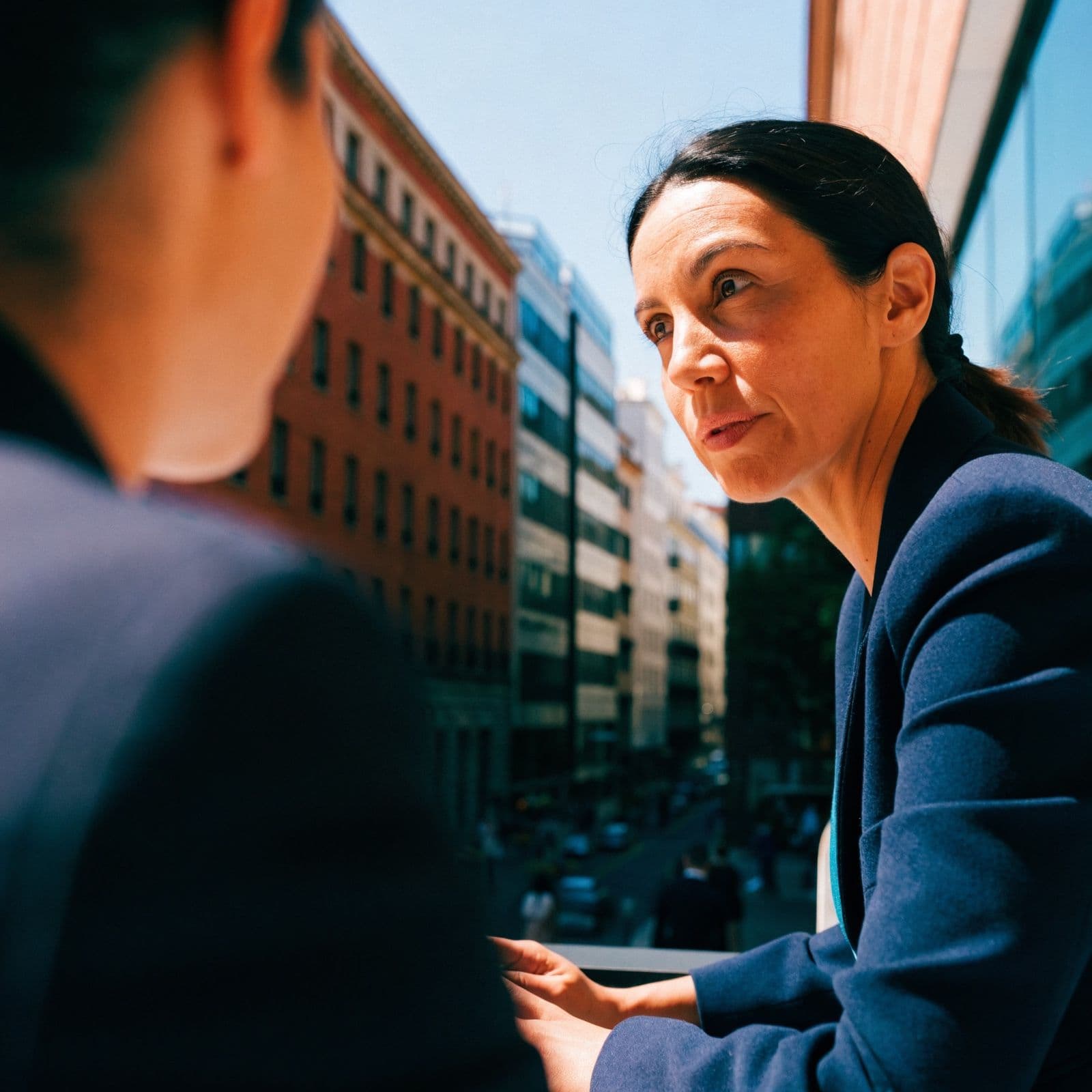 A leader leans in to question a colleague across a sunlit table, asking why this, why now and on what evidence.