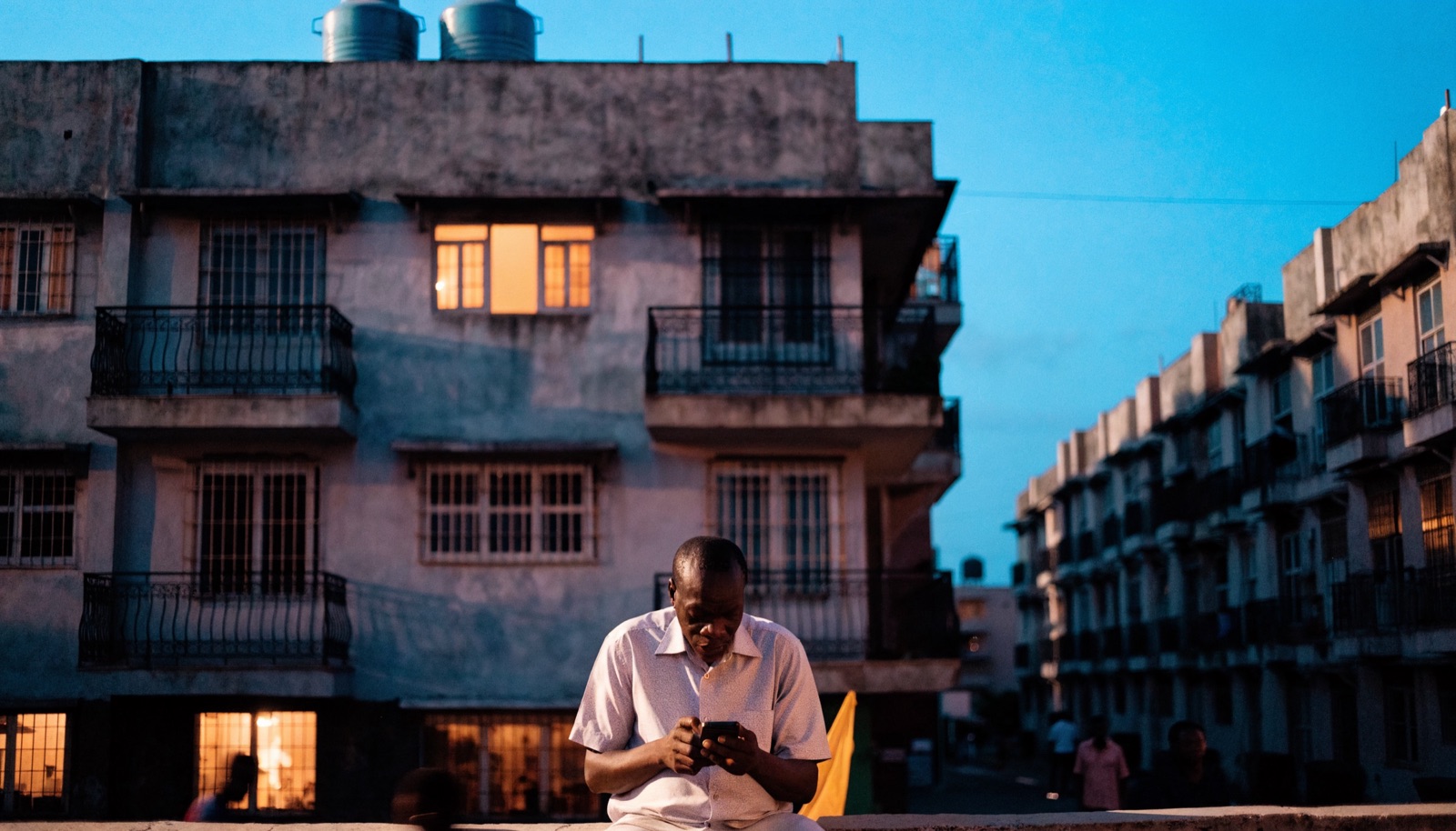 Illustration: a man outside a Lagos apartment block in the evening, reading his phone after work.