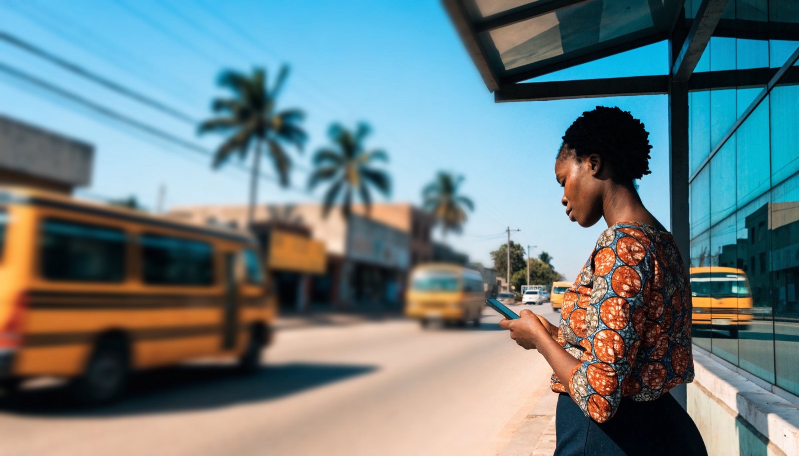 Illustration: a woman at a Lagos bus stop in the morning, glancing at her phone as minibuses pass.
